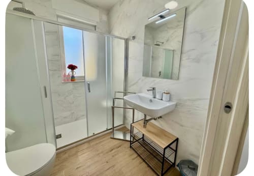 Bagno moderno nell'appartamento dell'aeroporto di Bari con pareti in marmo, doccia in vetro, lavabo bianco e pavimento in legno