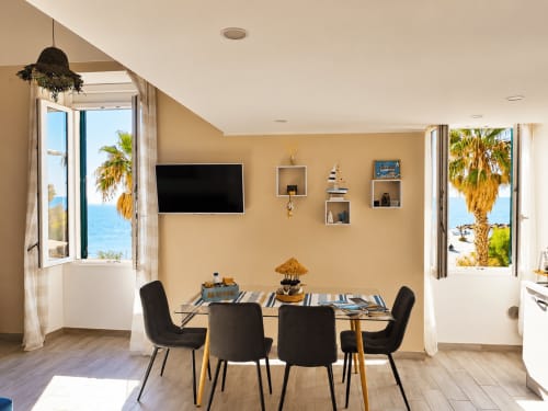 Elegante zona pranzo in appartamento beige con vista oceano, sedie nere e palme visibili attraverso le porte aperte a TuffonelBlu,