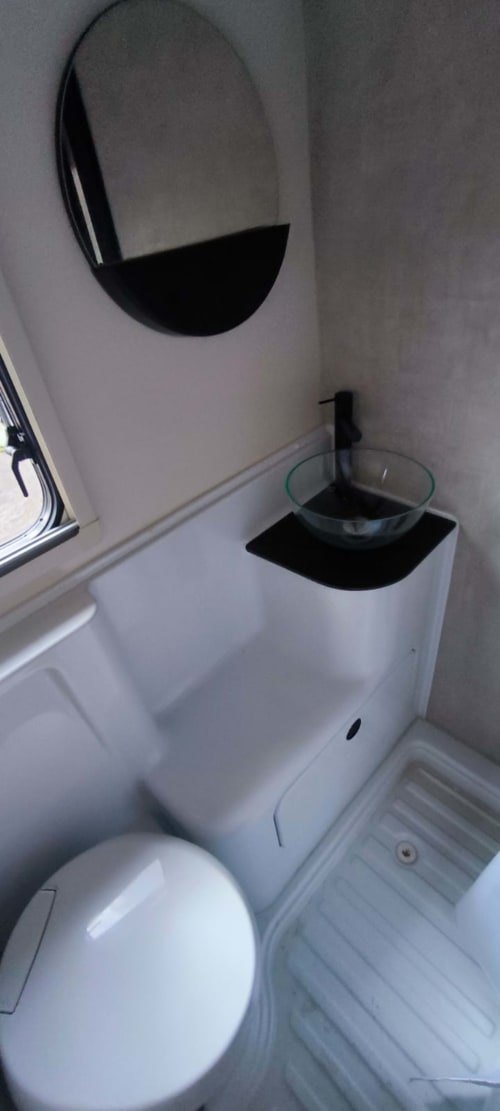 Baño moderno y compacto en la autocaravana Gozamena, Corralejo, con inodoro blanco, lavabo de cristal, espejo ovalado y pequeña ventana