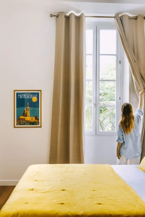 Chambre lumineuse du Boutique Hôtel Provencia avec lit jaune, rideaux beige et affiche de Marseille près de la fenêtre.