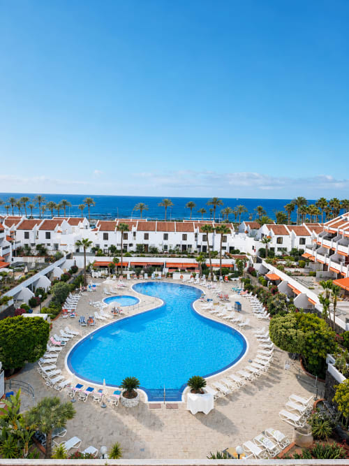 Aerial view of Playa de las Américas resort with blue pool, white apartments, palm trees and ocean in Tenerife
