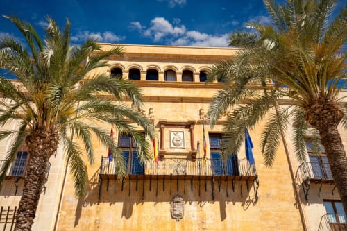 Hotel boutique Velarde en Elche con fachada de piedra amarilla, palmeras y ventanas arqueadas bajo cielo azul