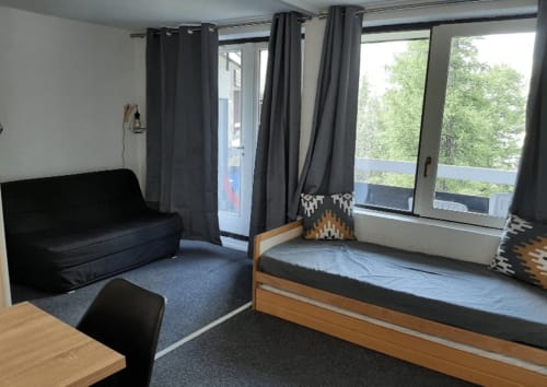Studio GNM à Vars : chambre avec lit en bois, canapé noir, grandes fenêtres sur la verdure et rideaux foncés