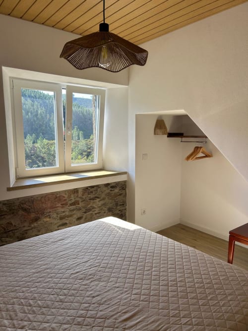Quarto luminoso com cama acolchoada, parede de pedra, teto de madeira, luminária pendente e vistas da montanha pelas grandes janelas da Casas