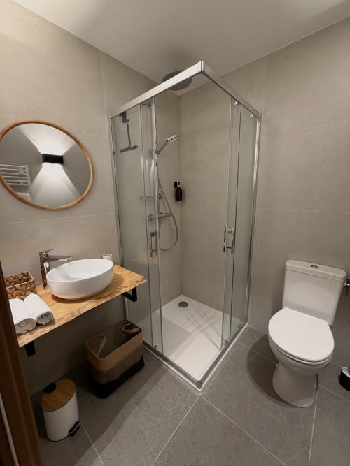 Salle de bains moderne de l'Hôtel les Vieilles Pierres avec douche vitrée, miroir rond, vasque en bois et toilettes