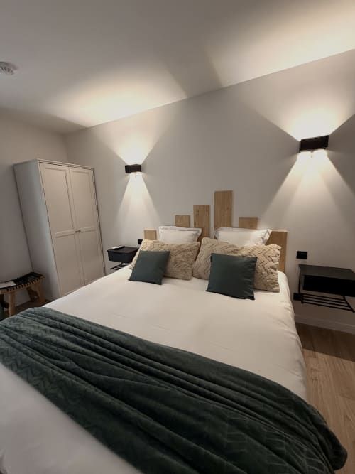 Chambre moderne à l'Hôtel les Vieilles Pierres avec draps blancs, tête de lit en bois et éclairage mural ambiant