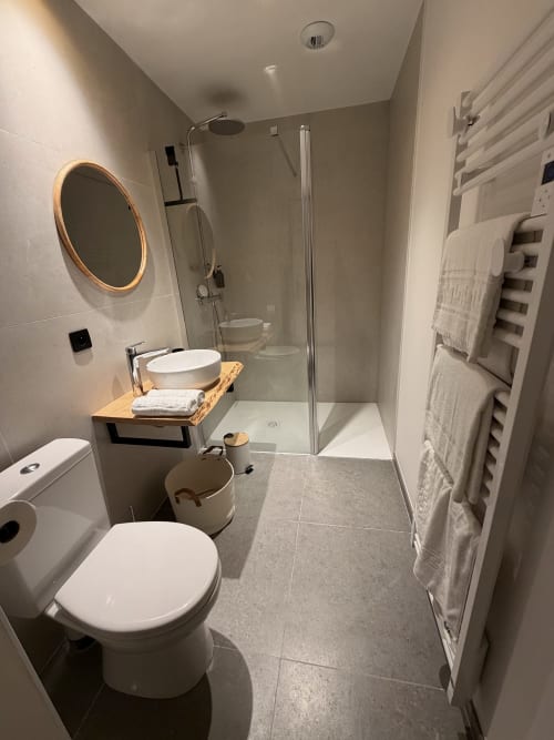 Salle de bain moderne à l'Hôtel les Vieilles Pierres à Eymet avec toilettes blanches, miroir rond, meuble vasque en bois et douche à l'italienne