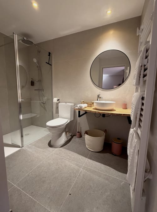 Salle de bain moderne à l'Hôtel les Vieilles Pierres avec douche à l'italienne, miroir rond, meuble vasque en bois et sèche-serviettes électrique
