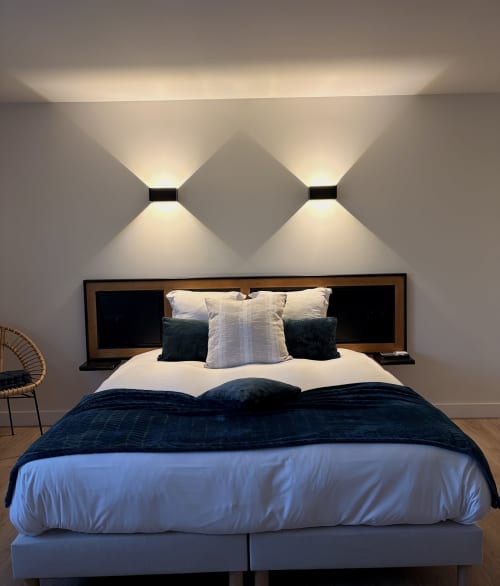 Chambre moderne à l'Hôtel les Vieilles Pierres avec éclairage mural géométrique, literie bleu foncé et tête de lit en bois
