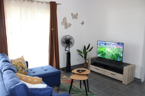 Salon moderne de Maisons En Or Cayenne avec canapé bleu, télévision, ventilateur et vue sur le jardin