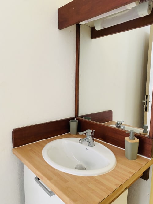 Salle de bains moderne avec lavabo blanc rond, plan de travail en bois et encadrement en bois foncé à l'appartement Maisons En Or Sinnamary