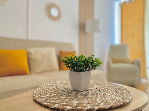 Salon moderne Maisons En Or avec plante en pot sur table tissée, canapé crème aux coussins jaunes et porte jaune