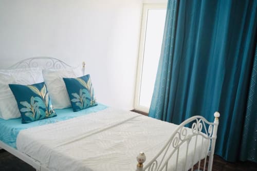 Chambre lumineuse avec lit en métal blanc, coussins turquoise et rideaux bleu-vert aux Maisons En Or à Cayenne