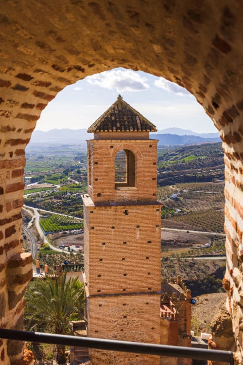 Torre alminar de ladrillo rojo vista a través de un arco que domina el valle de Álora y los olivares de Andalucía, España