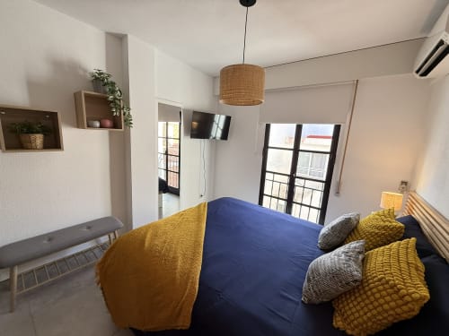 Dormitorio moderno en apartamento de Fuengirola con cama azul, detalles amarillos, TV, balcón y vistas a la ciudad