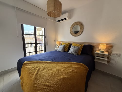 Moderno dormitorio en apartamento de Fuengirola con cama azul, detalles dorados, cabecero de madera, balcón con vistas a la ciudad y