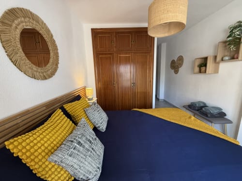 Dormitorio moderno en apartamento de Fuengirola con armario de madera, ropa de cama amarilla, cama azul marino e iluminación natural de lámpara colgante