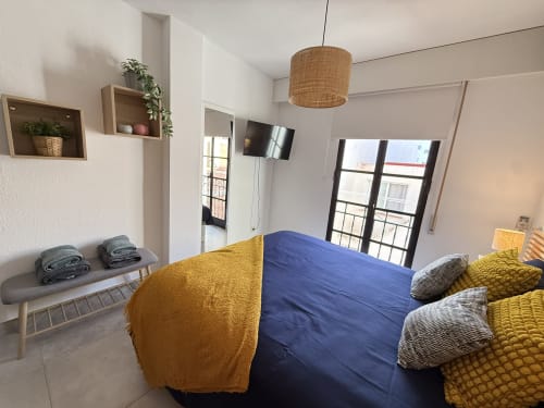 Dormitorio moderno en apartamento de Fuengirola con cama azul, detalles amarillos, TV de pantalla plana, balcón con vista a la ciudad
