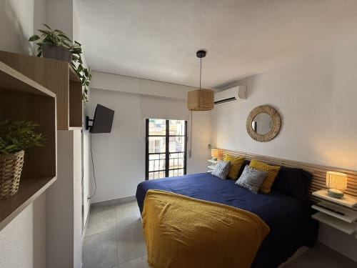 Dormitorio moderno en apartamento de Fuengirola con cama azul, detalles amarillos, vista al balcón, lámpara colgante de ratán y televisor.
