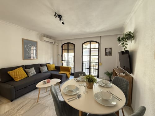 Moderna sala de estar en Fuengirola con mesa de comedor, sofá negro, detalles amarillos y acceso a balcón