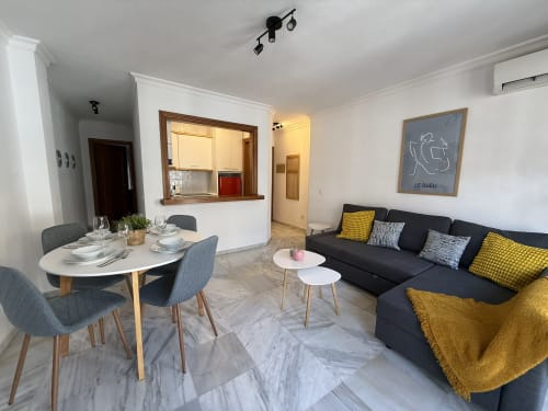 Moderno salón de apartamento en Fuengirola con sofá negro, mesa de comedor, cocina y detalles en amarillo