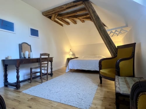 Chambre rustique avec lit à baldaquin en bois, poutres apparentes, luminosité chaleureuse et fauteuil vert vintage aux Anciennes