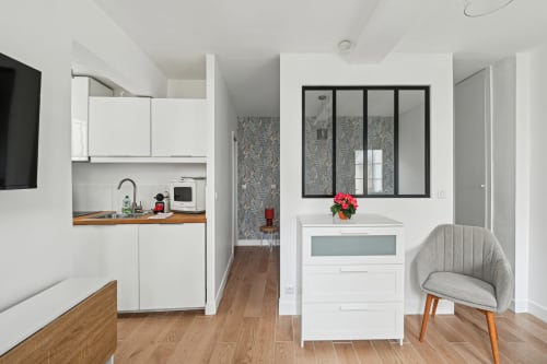 Kitchenette moderne avec armoires blanches, comptoirs en bois et salon avec fauteuil gris à la Résidence Aurmat Paris