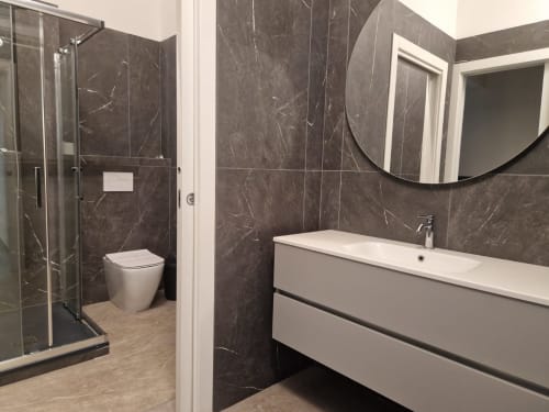 Bagno moderno nell'appartamento Fresia House a Genova. Piastrelle in marmo scuro, lavabo bianco, specchio curvo e doccia chiusa