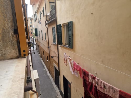 Stretto vicolo genovese con edifici in cotto, bucato rosso steso, persiane verdi, appartamento con vista sulla città