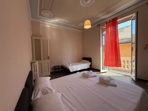 Ampia camera da letto nell'appartamento Tulipano di ML White House a Genova con due letti, balcone con vista sulla città e tende rosse.