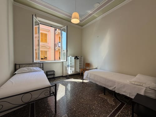 Camera luminosa a Tulipano House a Genova con due letti, lampada a sospensione, finestra aperta con vista sulla città, pavimento in terrazzo