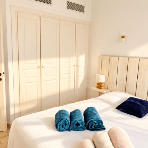 Chambre lumineuse aux Villas d'A Storia à Lecci avec serviettes roulées sur le lit, tête de lit en bois, armoire crème et lumière naturelle