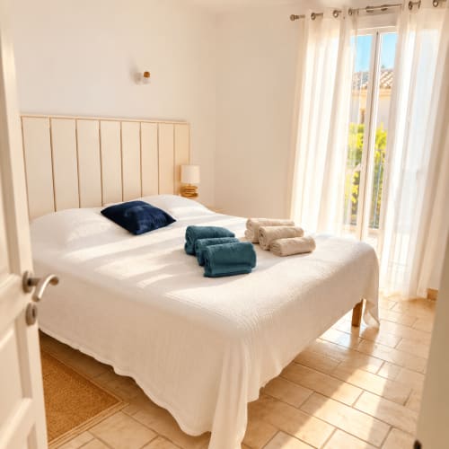 Chambre lumineuse aux Villas d'A Storia à Lecci avec lit blanc, tête de lit en bois, oreiller bleu, serviettes roulées et vitre