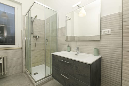 Moderno bagno nell'appartamento ERICA HOUSE a Genova. Piastrelle grigie, vanità scura, lavabo bianco, box doccia in vetro