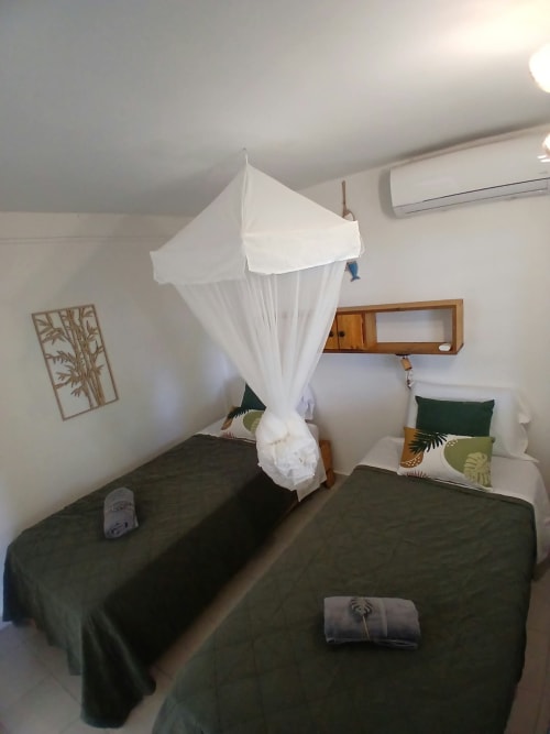 Chambre tropicale avec moustiquaire blanche en baldaquin, lits jumeaux aux housses vert olive, étagères en bois et climatiseur au plafond