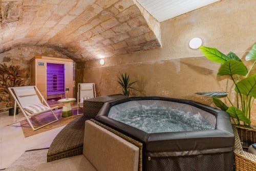 Suite spa luxueuse Wellkhome Bordeaux avec jacuzzi, sauna infrarouge, chaise longue et plantes verdoyantes dans une pièce aux murs de pierre