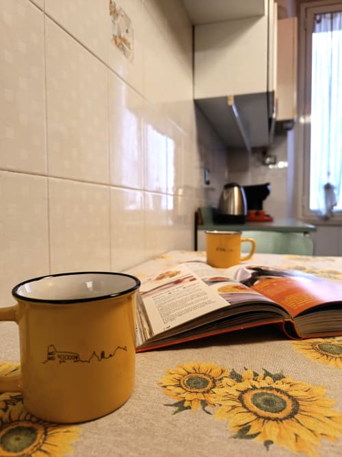 Tavolo accogliente della cucina a Sweet Home Rome con tazza gialla, rivista aperta e tessuto a motivi di girasoli nell'appartamento Matilde
