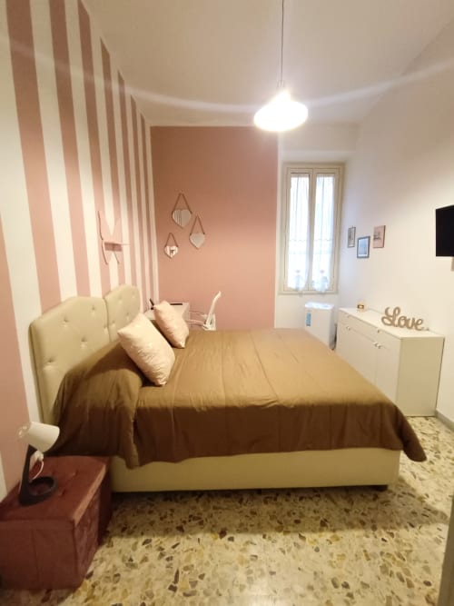 Camera da letto Matilde Sweet Home a Roma con parete a righe rosa, letto color crema, comodino in legno e lampada a sospensione moderna