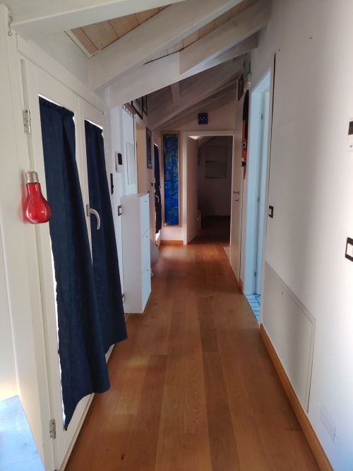 Lungo corridoio in appartamento di villa a Oristano con pavimenti in legno, pareti bianche, porte blu e luce di emergenza rossa