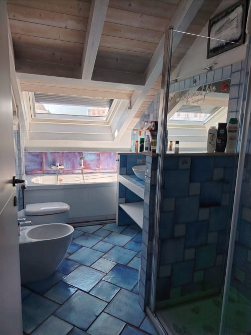 Bagno mansardato luminoso a Maison Belle Époque, Oristano con lucernario, pavimento in piastrelle blu, sanitari bianchi e vista sulla città.