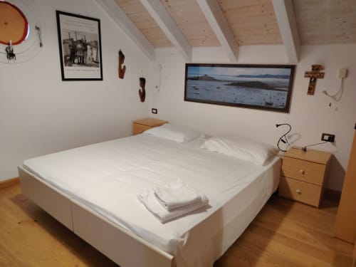 Camera mansardata luminosa con travi in legno, letto bianco con asciugamani, comodino e quadro di paesaggio costiero presso Maison Belle