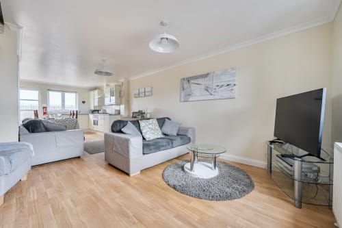 Open-plan living area in Exeter cottage with cream walls, wooden floors, gray sofas, flat-screen TV, and kitchen in
