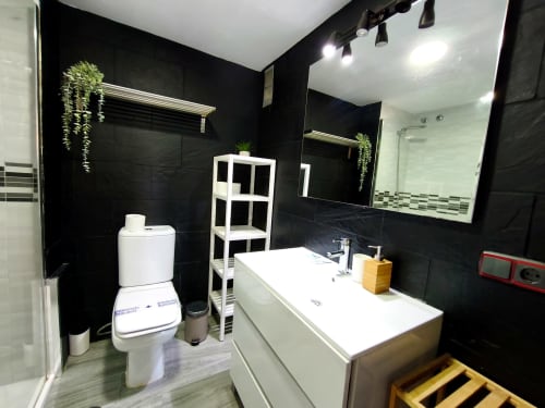 Baño moderno en blanco y negro con lavabo, inodoro, espejo y estanterías blancas en el apartamento VIVE Benalmádena