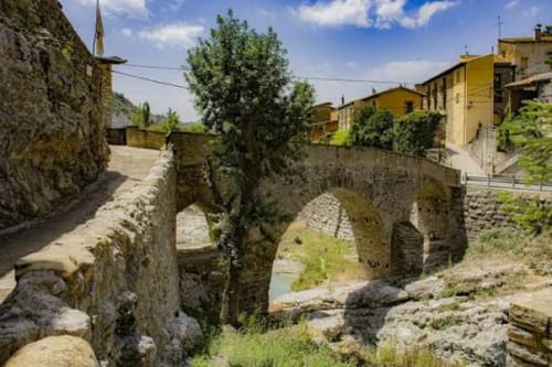 Puente medieval de piedra sobre arroyo en el pueblo pirenaico de Montgat con casas amarillas y vistas a las montañas