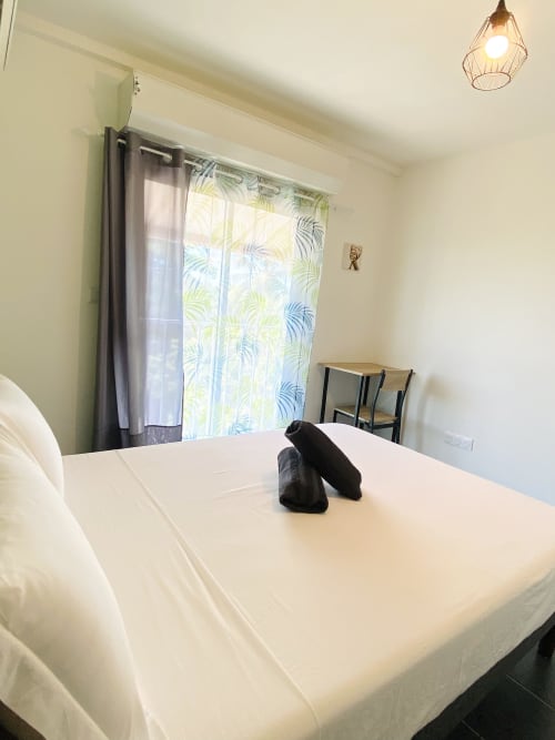 Chambre lumineuse et moderne dans l'appartement de Cayenne avec lit blanc, rideaux motifs tropicaux et vue sur mer aux Maisons En Or
