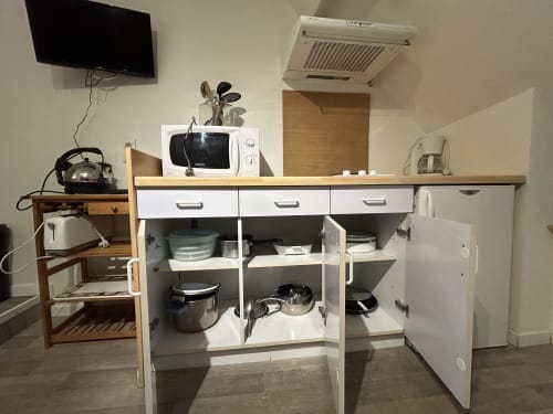 Kitchenette compacte du Studio Hébergement Luxeuil Les Bains avec micro-ondes, réfrigérateur, ustensiles de cuisine et TV.