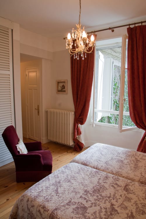 Chambre élégante aux Chambres d'hôtes Les Pratges à Figeac avec lits jumeaux, fauteuil bordeaux, lustre et portes-fenêtres