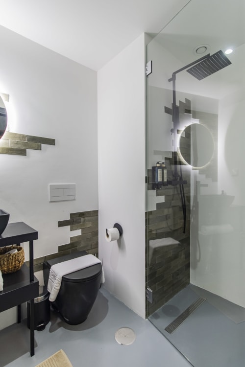 Banheiro moderno em hotel no Porto com vaso sanitário preto, parede de destaque em azulejos escuros, chuveiro tipo chuva, espelho circular iluminado e