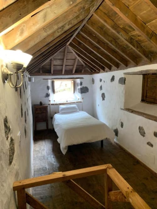 Dormitorio rústico en buhardilla de Casa 1820, La Palma rural, con vigas de madera, paredes blancas, cama individual y ventana con vistas al jardín