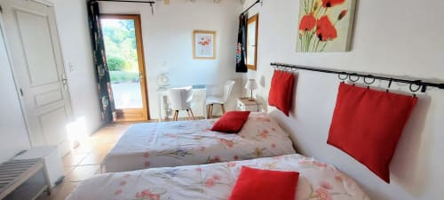 Bright cottage bedroom with two beds featuring floral bedding, red pillows, garden views, and modern amenities at Les Gites